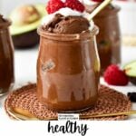 a glass jar filled with Chocolate Avocado Mousse topped with whipped cream, raspberries and a golden spoon digging in the mousse showing its airy texture.