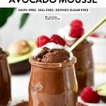 a glass jar filled with Chocolate Avocado Mousse topped with whipped cream, raspberries and a golden spoon digging in the mousse showing its airy texture.