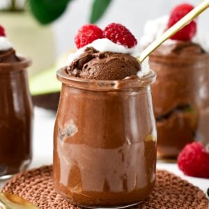 a glass jar filled with Chocolate Avocado Mousse topped with whipped cream, raspberries and a golden spoon digging in the mousse showing its airy texture.