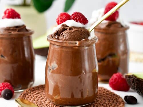 a glass jar filled with Chocolate Avocado Mousse topped with whipped cream, raspberries and a golden spoon digging in the mousse showing its airy texture.