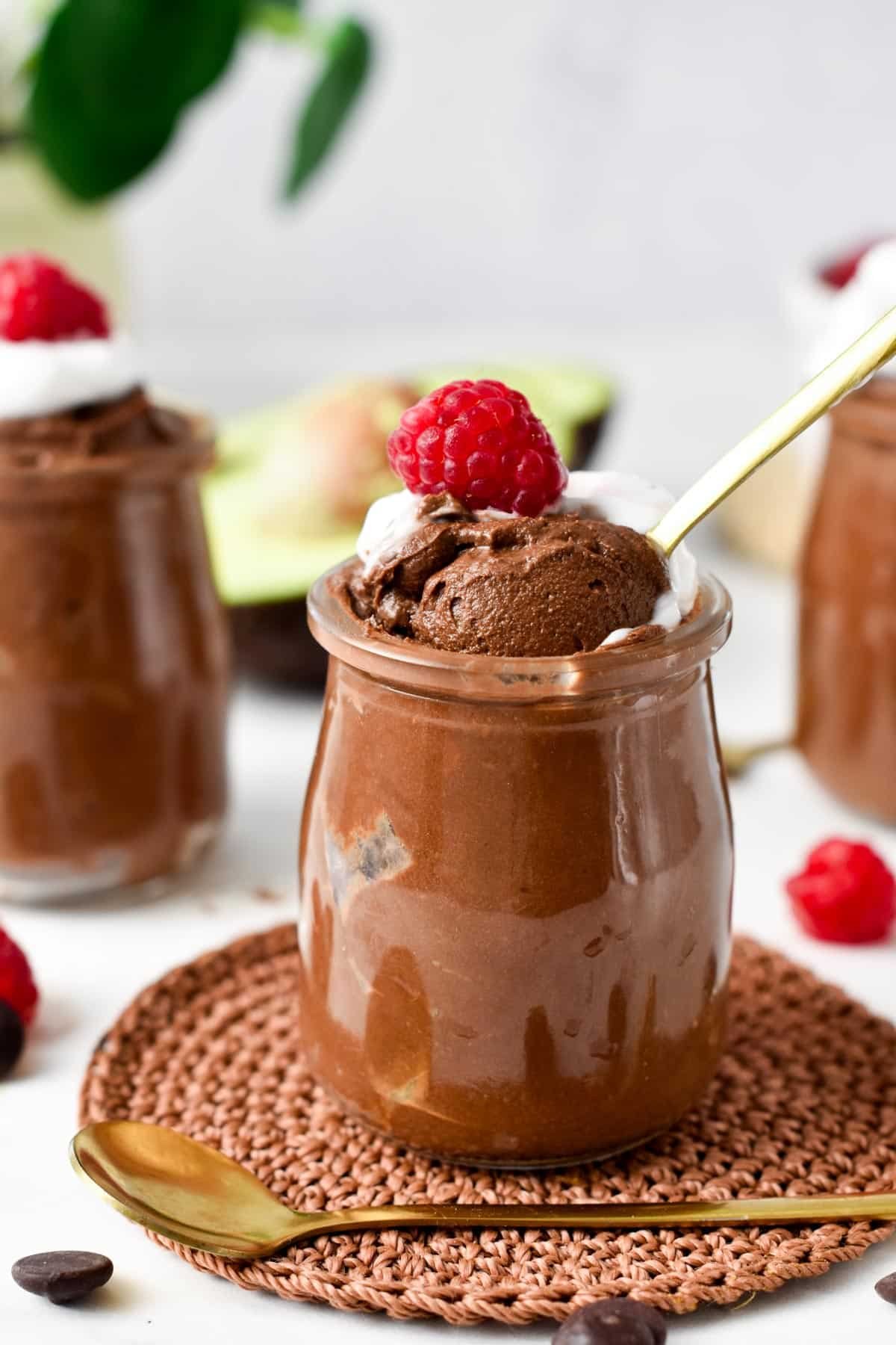 A glass jar filled with Chocolate Avocado Mousse topped with whipped cream, raspberries and a golden spoon digging in the mousse showing its airy texture. 