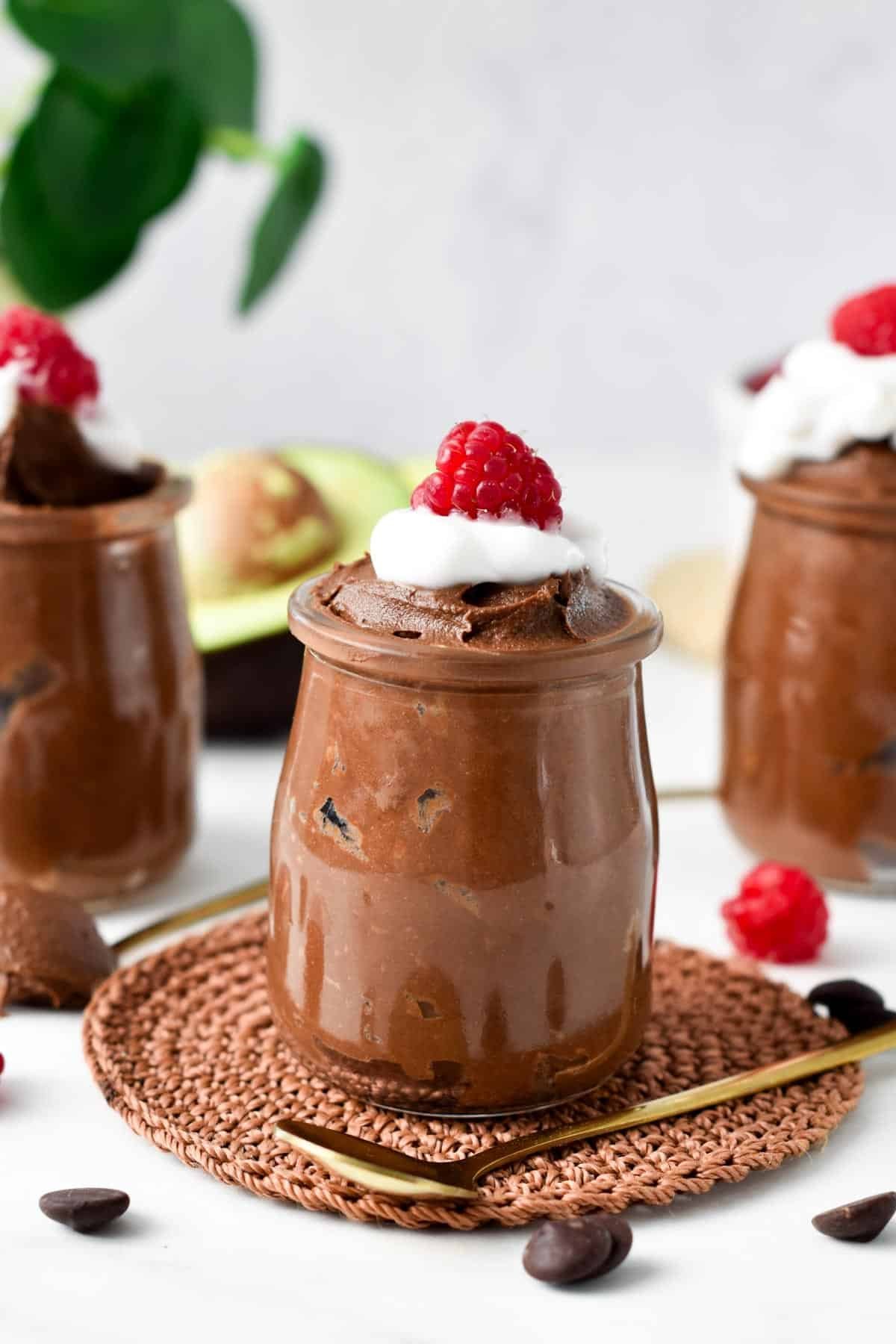 A glass jar filled with Chocolate Avocado Mousse topped with whipped cream, raspberries and avocado in the background.