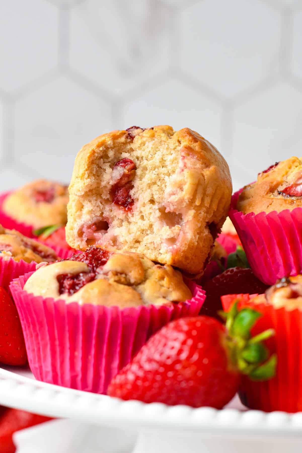 A stack of vegan strawberry muffins with a golden fluffy crumb filled with juicy pieces of baked strawberries and the top one half open, showing it's crumbs.