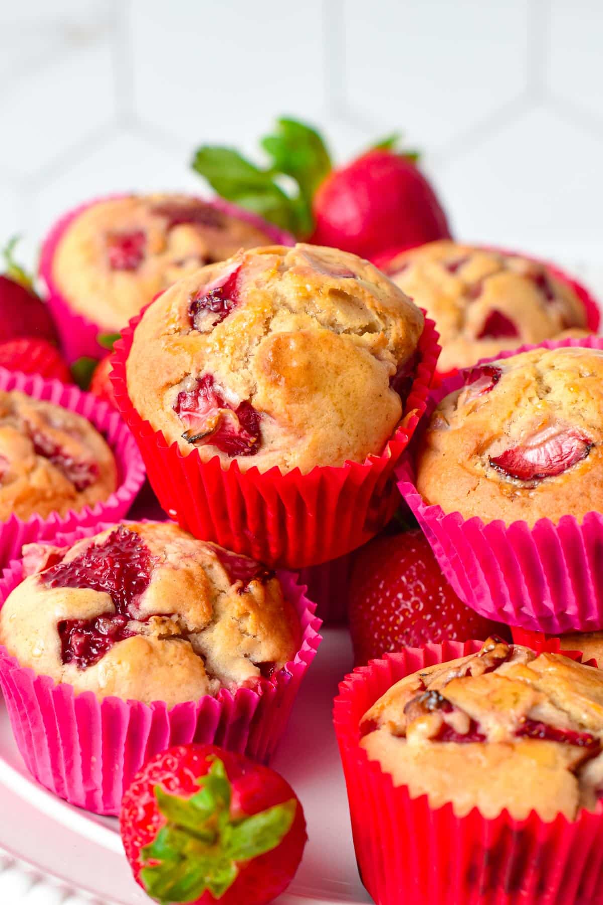 A stack of vegan strawberry muffins in bright pink muffin cases with a golden fluffy crumb filled with juicy pieces of baked strawberries.