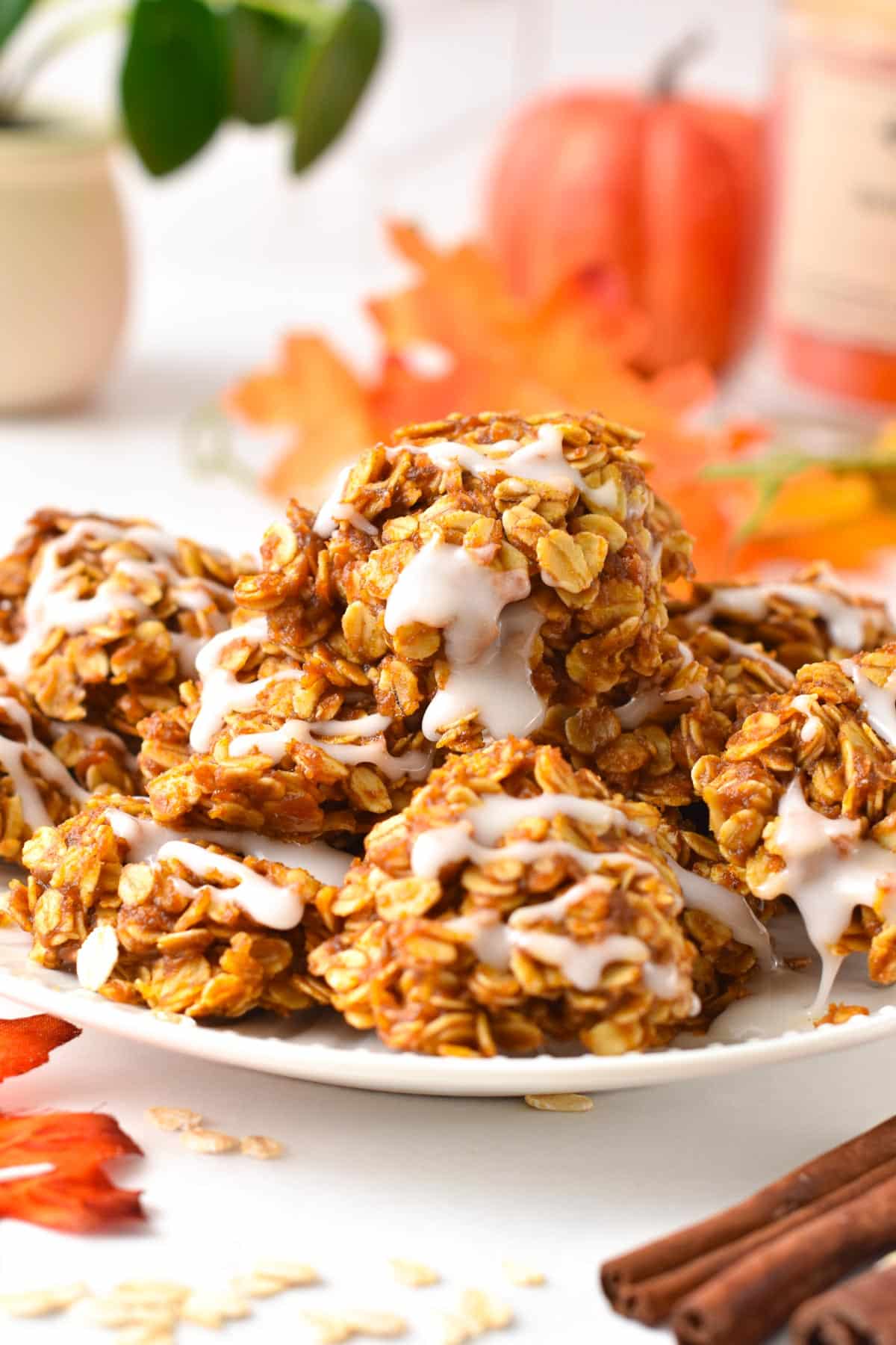 A plate filled with a stack of no-bake pumpkin cookies topped with a drizzle of icing sugar next to cinnamon sticks.