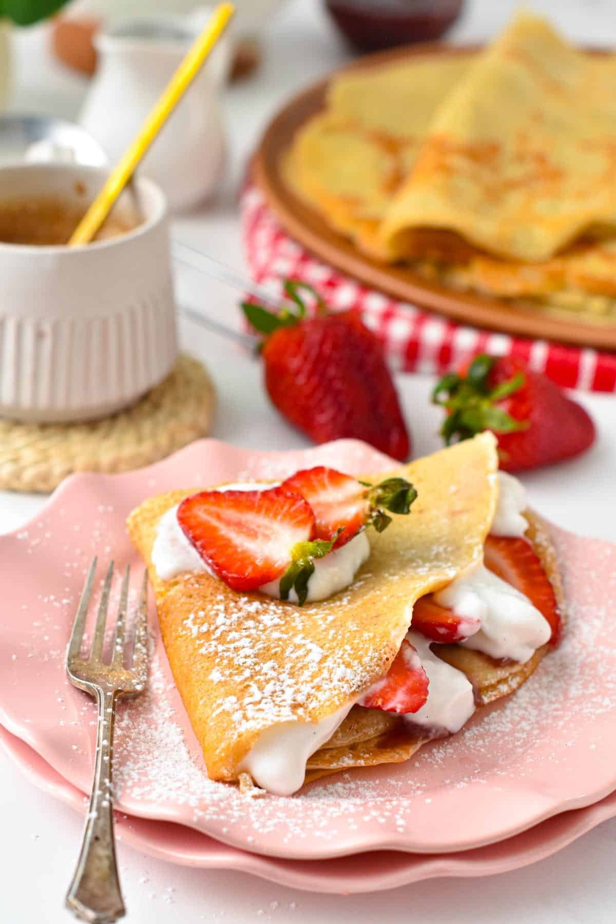 A vegan French crepe filled with strawberries, dairy-free whipped cream with more vegan crepes in the background.