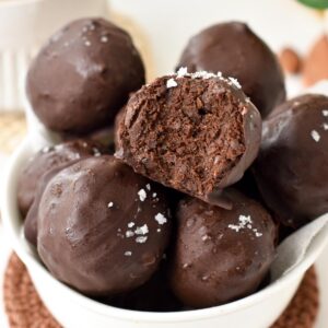a bowl filled with no-bake brownie protein balls with one ball showing the inside chocolate fudge texture