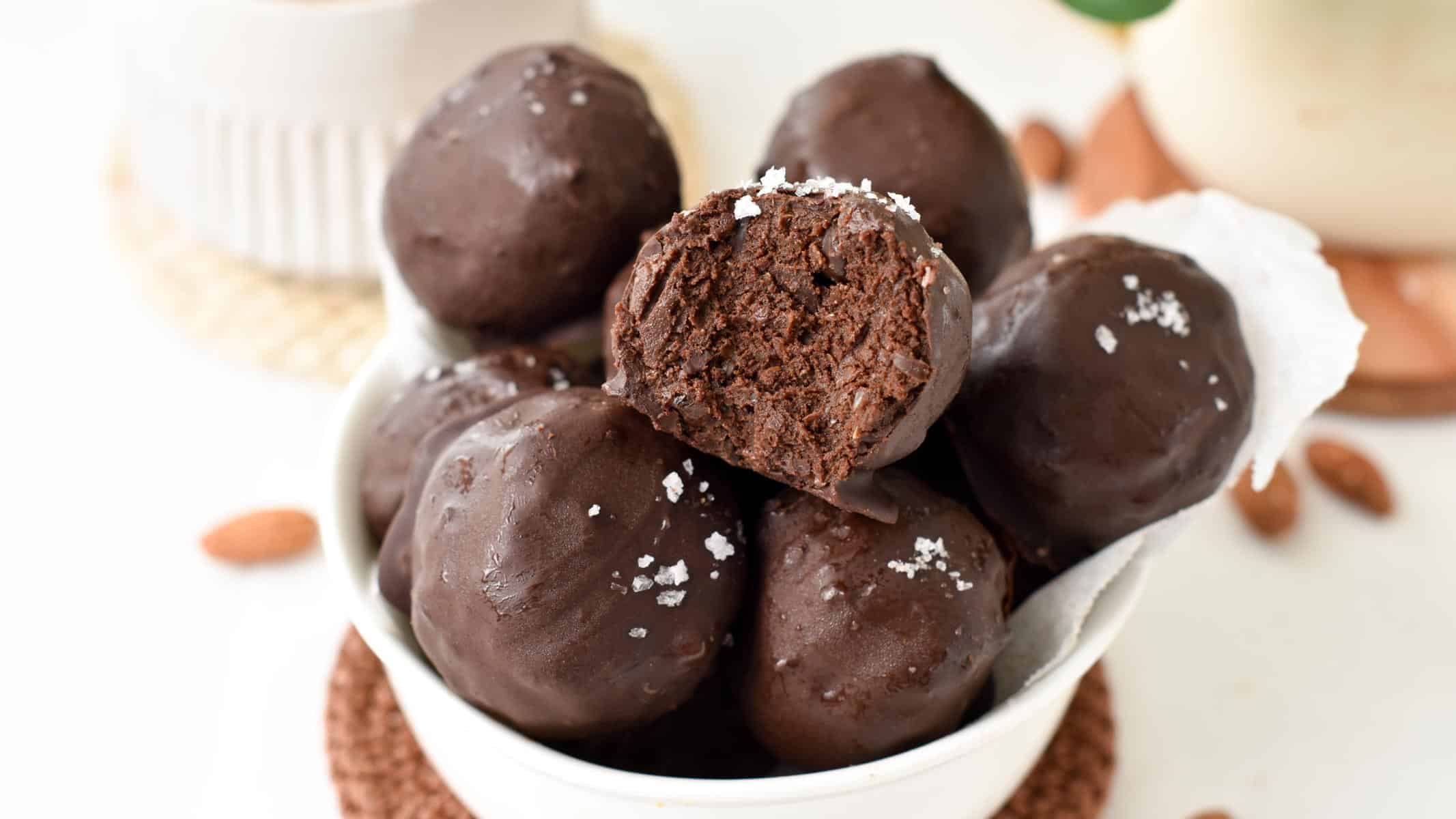 a bowl filled with no-bake brownie protein balls with one ball showing the inside chocolate fudge texture