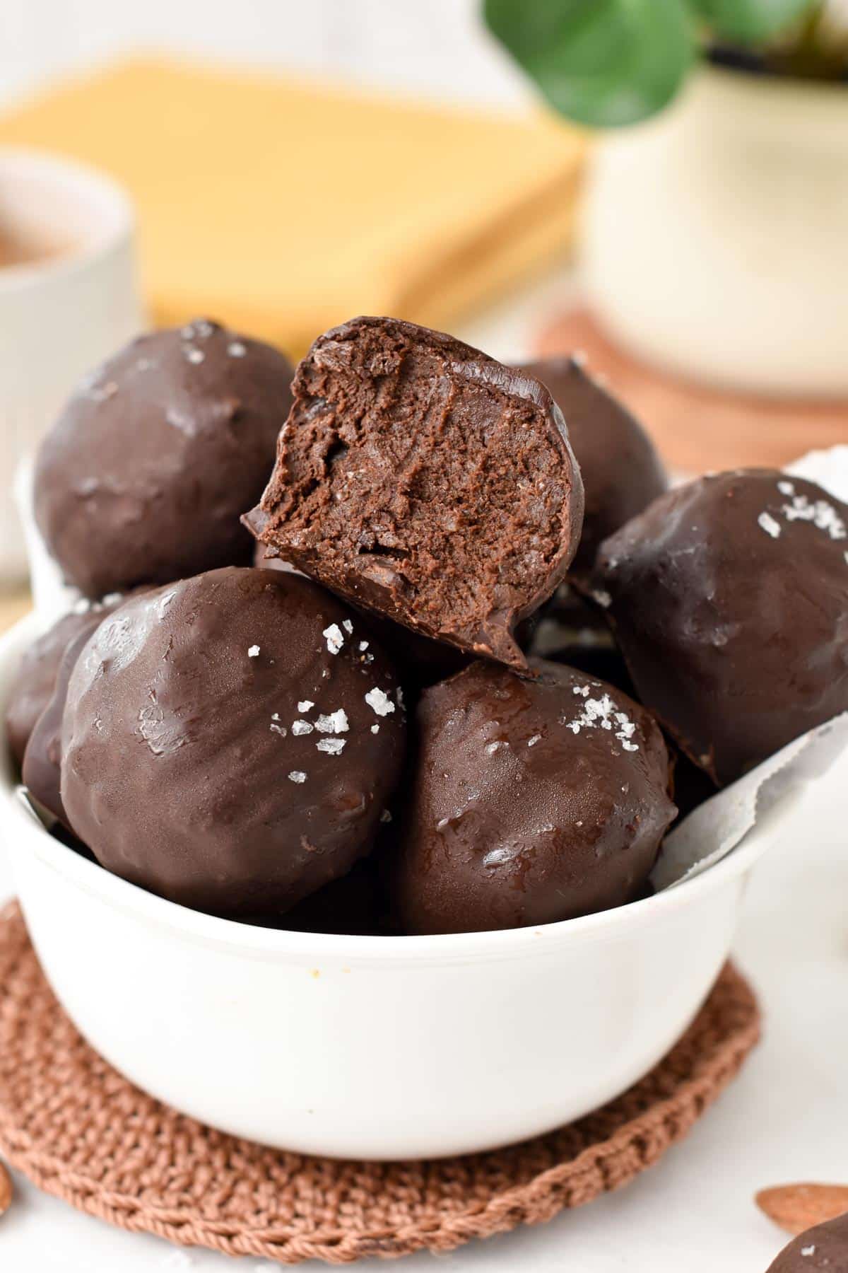 A bowl filled with no-bake brownie protein balls with one ball showing the inside chocolate fudge texture.