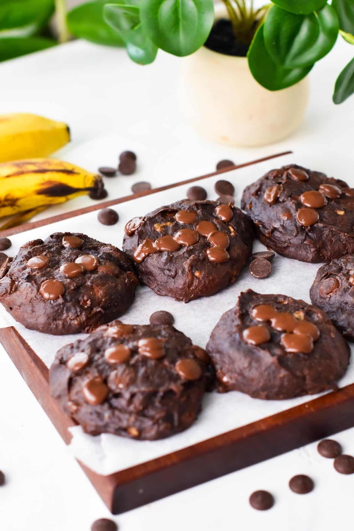 Six Chocolate Banana Cookies next to each other on a chopping board covered with white parchment paper.