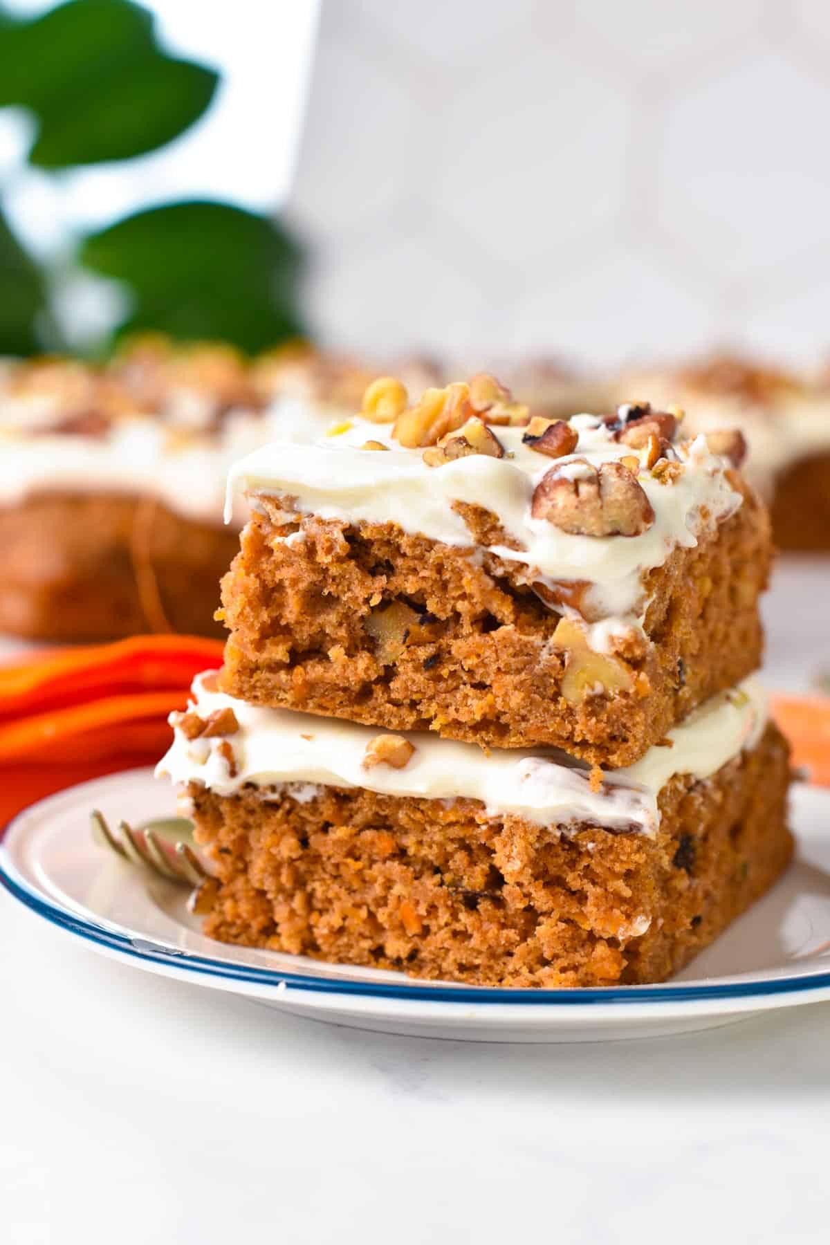 Two slices of sheet pan carrot cake, frosted, and stacked on a plate with in the background a carrot cake and green plant.