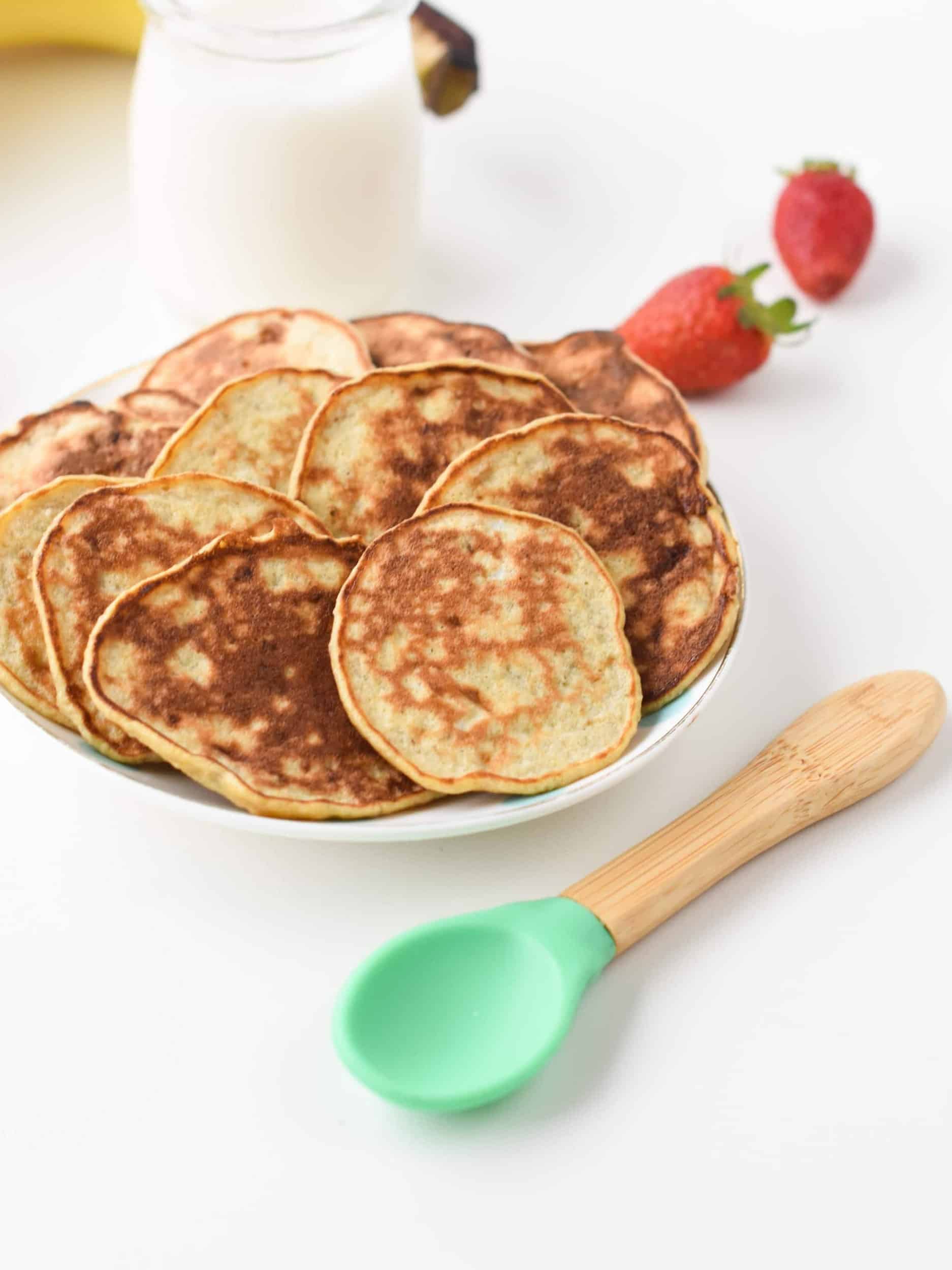 Baby Banana Pancakes on a small plate next to a kid's spoon and a glass of plant-based milk and strawberries.