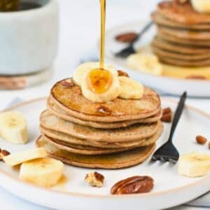 3-Ingredient Banana Oat Pancakes served on a white plate with a black fork and decorated with banana slices, pecan nuts, and maple syrup.