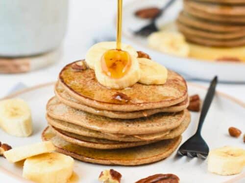 3-Ingredient Banana Oat Pancakes served on a white plate with a black fork and decorated with banana slices, pecan nuts, and maple syrup.