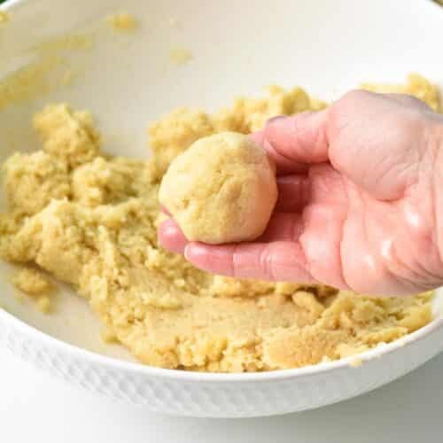 A hand holding a ball of almond flour banana cookie dough with in the background a mixing bowl with cookie dough.
