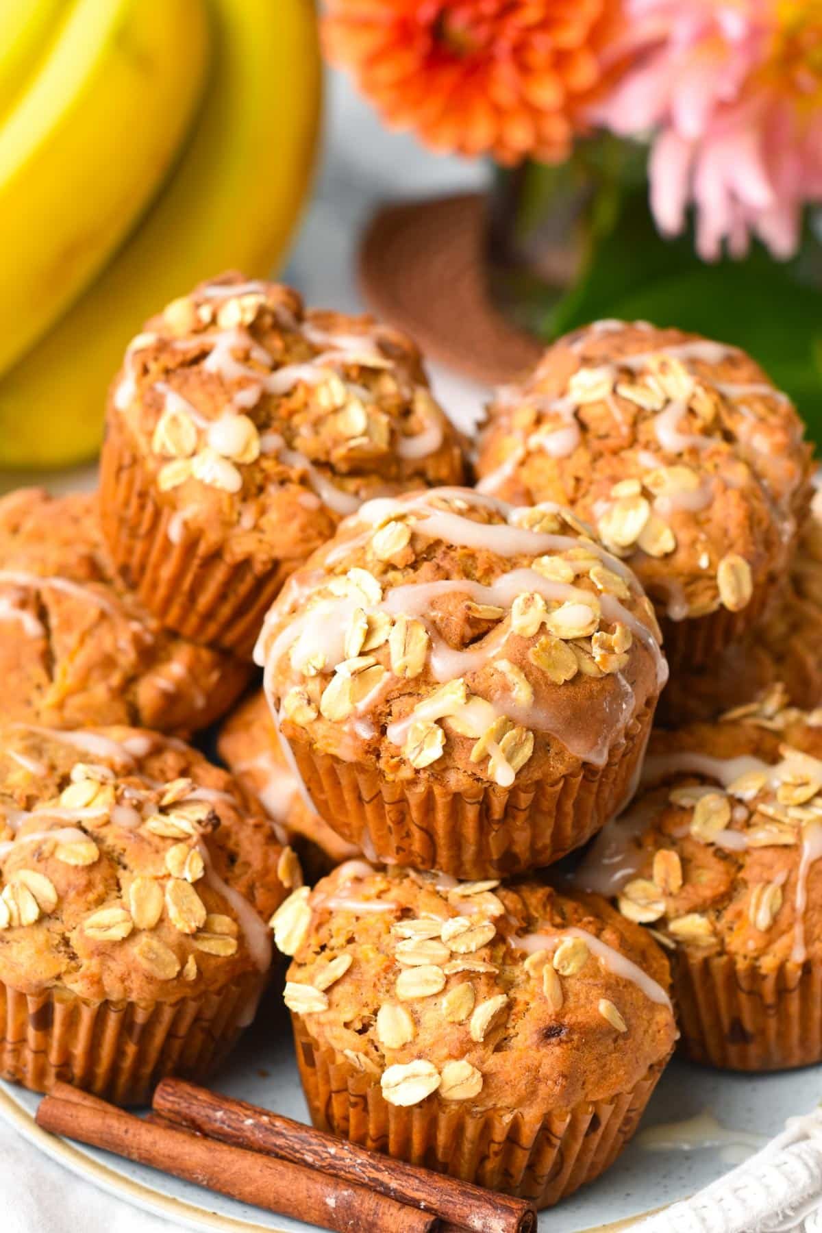 A plate filled with a stack of banana carrot muffins with oats on op and banana and orange flowers in the background.