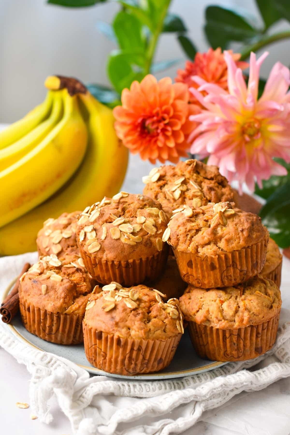 A plate filled with a stack of banana carrot muffins with oats on op and banana and orange flowers in the background