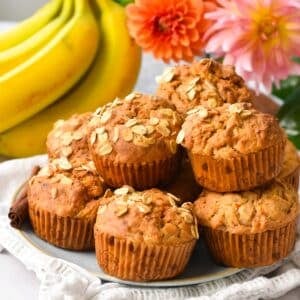 a plate filled with a stack of banana carrot muffins with oats on op and banana and orange flowers in the background