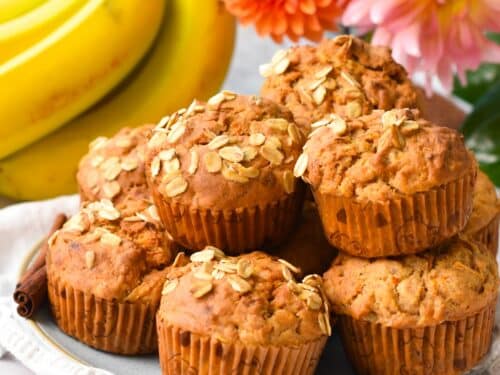 a plate filled with a stack of banana carrot muffins with oats on op and banana and orange flowers in the background