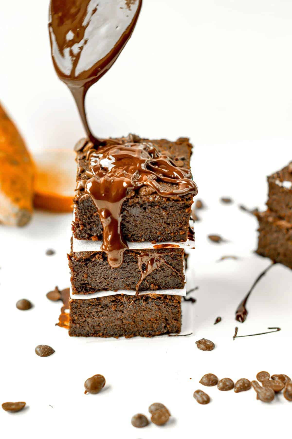 Vegan Sweet Potato Brownies stacked on a white table with melted chocolate dripping from a spoon.