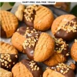 a black plate lined with white parchment paper and filled with lots of peanut butter cookies, half way dipped in dark chocolate and covered with crushed peanuts