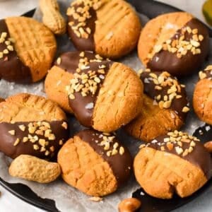 a black plate lined with white parchment paper and filled with lots of peanut butter cookies, half way dipped in dark chocolate and covered with crushed peanuts