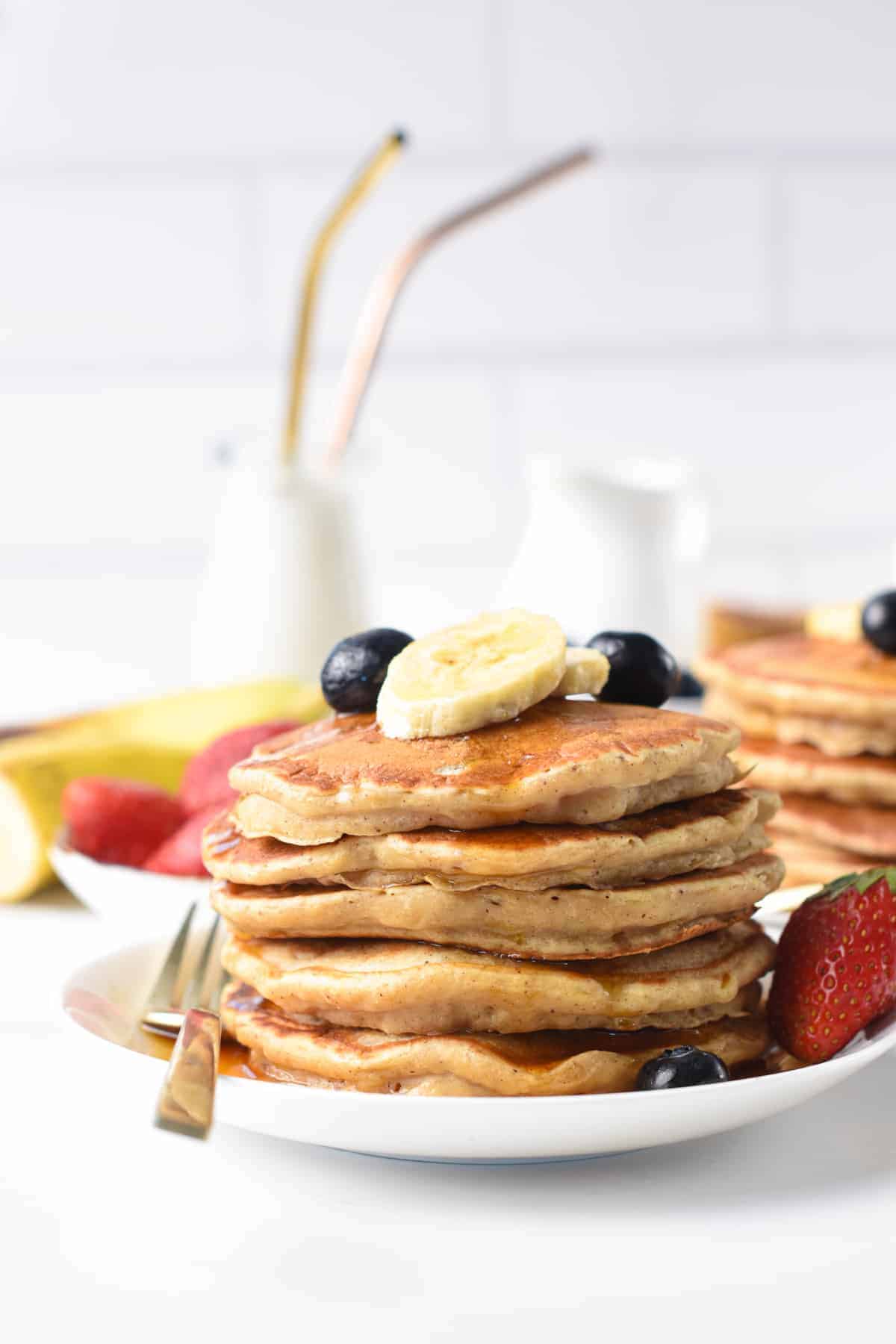 Vegan Banana Pancakes stacked on a white plate and decorated with fresh berries and bananas.