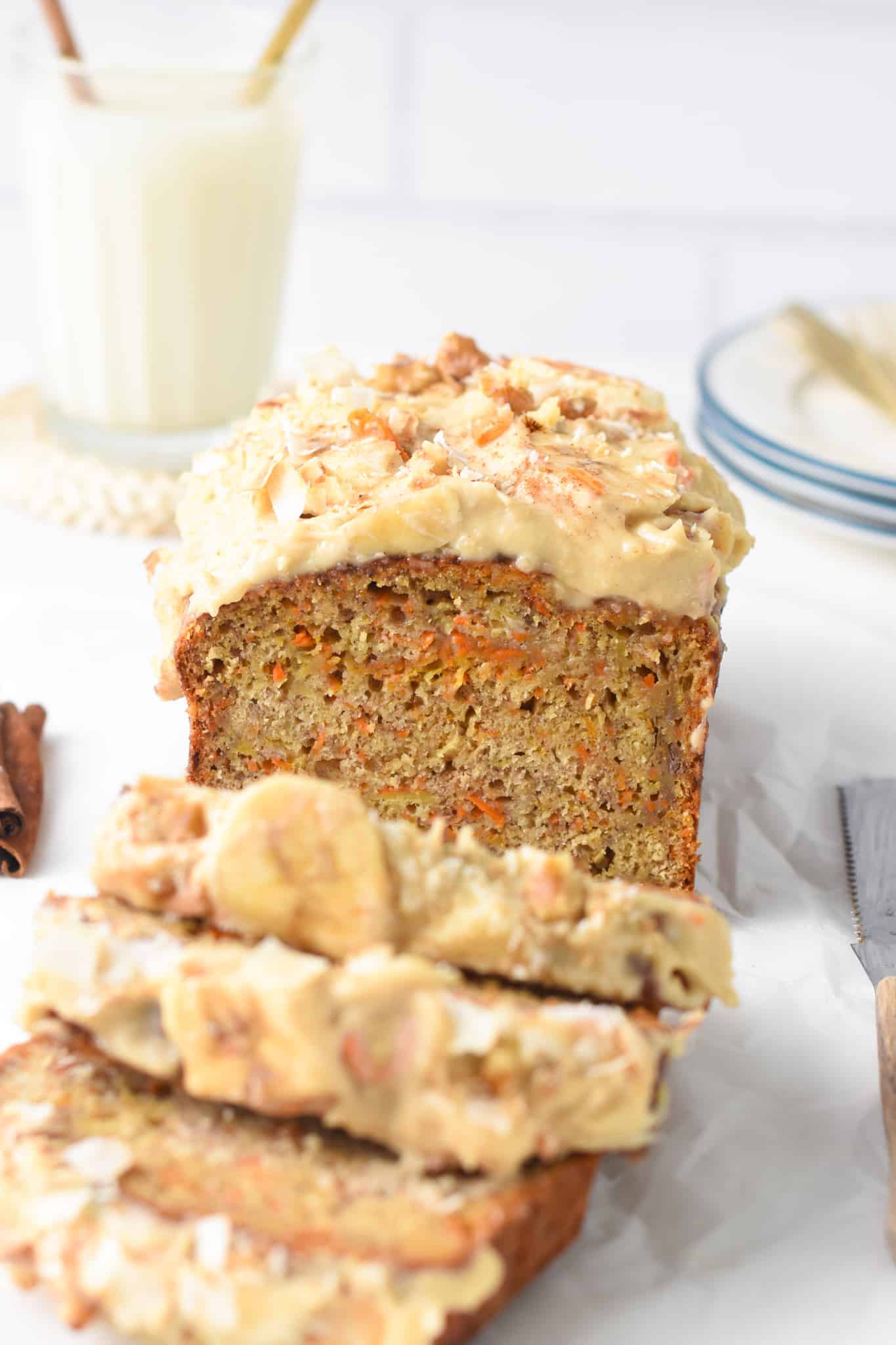 Carrot Banana Bread sliced in front of a glass of almond milk and a stack of dessert plates.