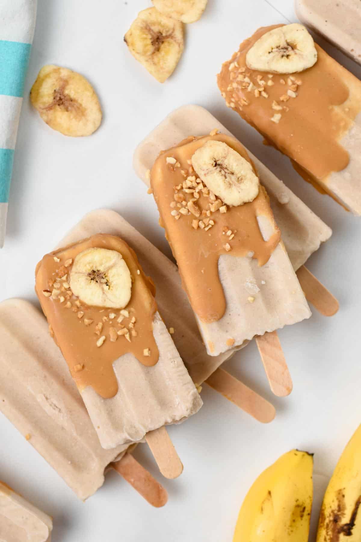 Banana Popsicles dipped in peanut butter and decorated with chopped nuts and banana and stacked on a table.