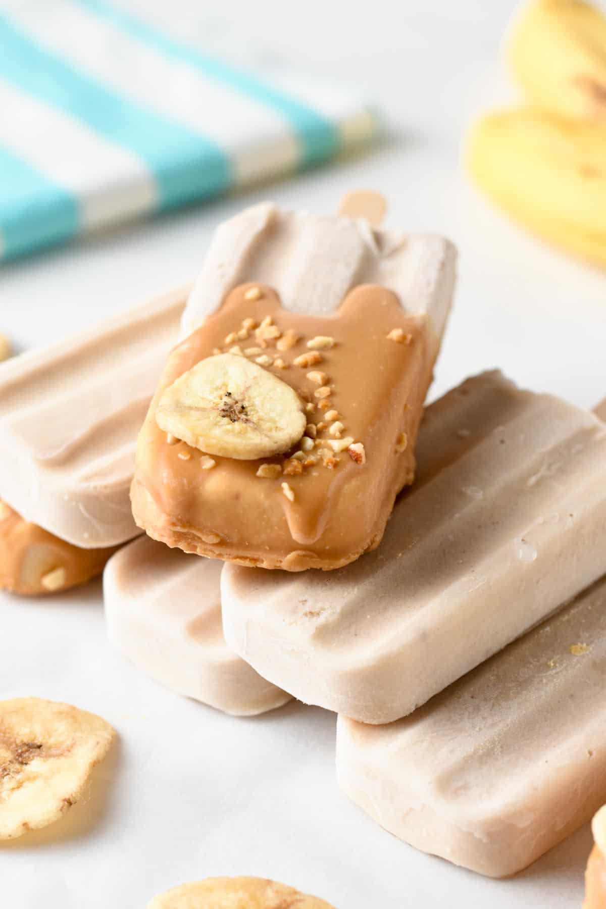 Banana Popsicles dipped in peanut butter and decorated with chopped nuts and banana and stacked on a table.