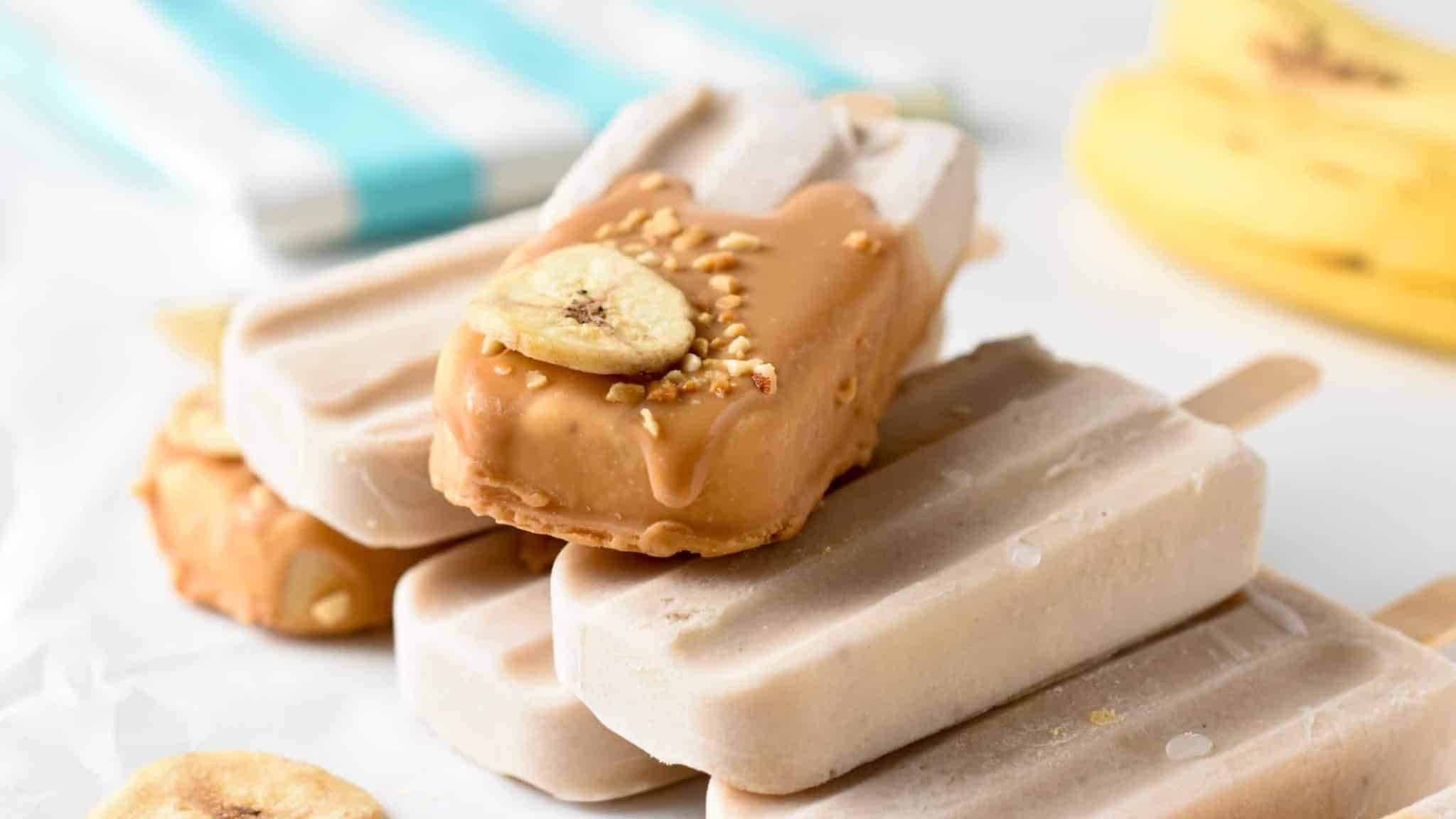 Banana Popsicles dipped in peanut butter and decorated with chopped nuts and banana and stacked on a table.