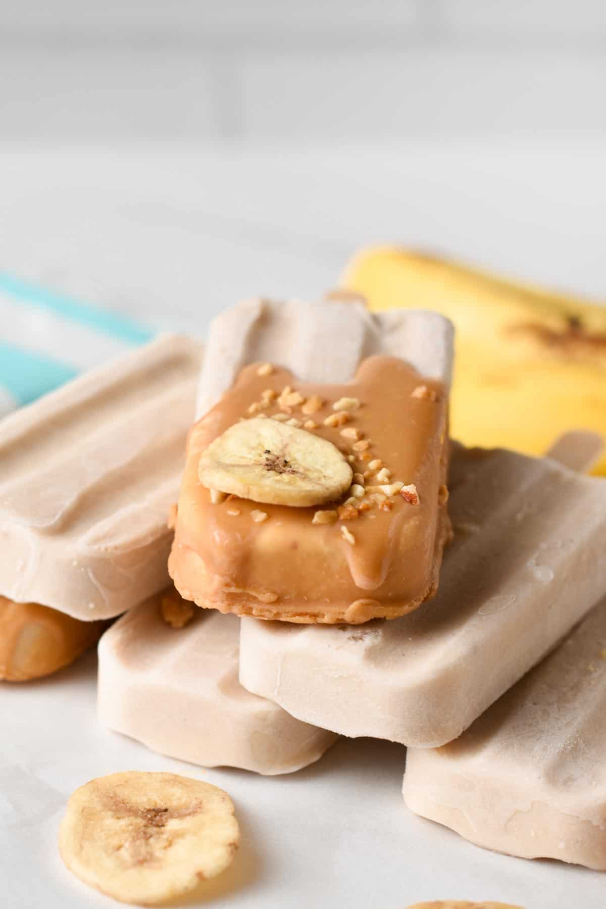 Banana Popsicles dipped in peanut butter and decorated with chopped nuts and banana and stacked on a table.