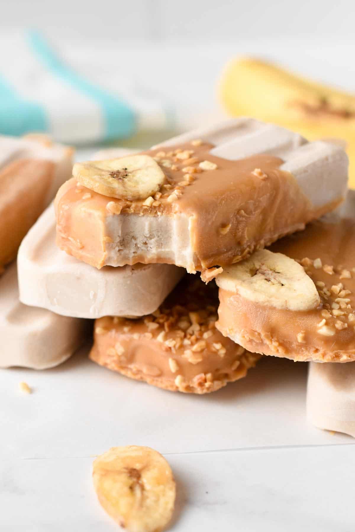 Banana Popsicles dipped in peanut butter and decorated with chopped nuts and banana and stacked on a table.