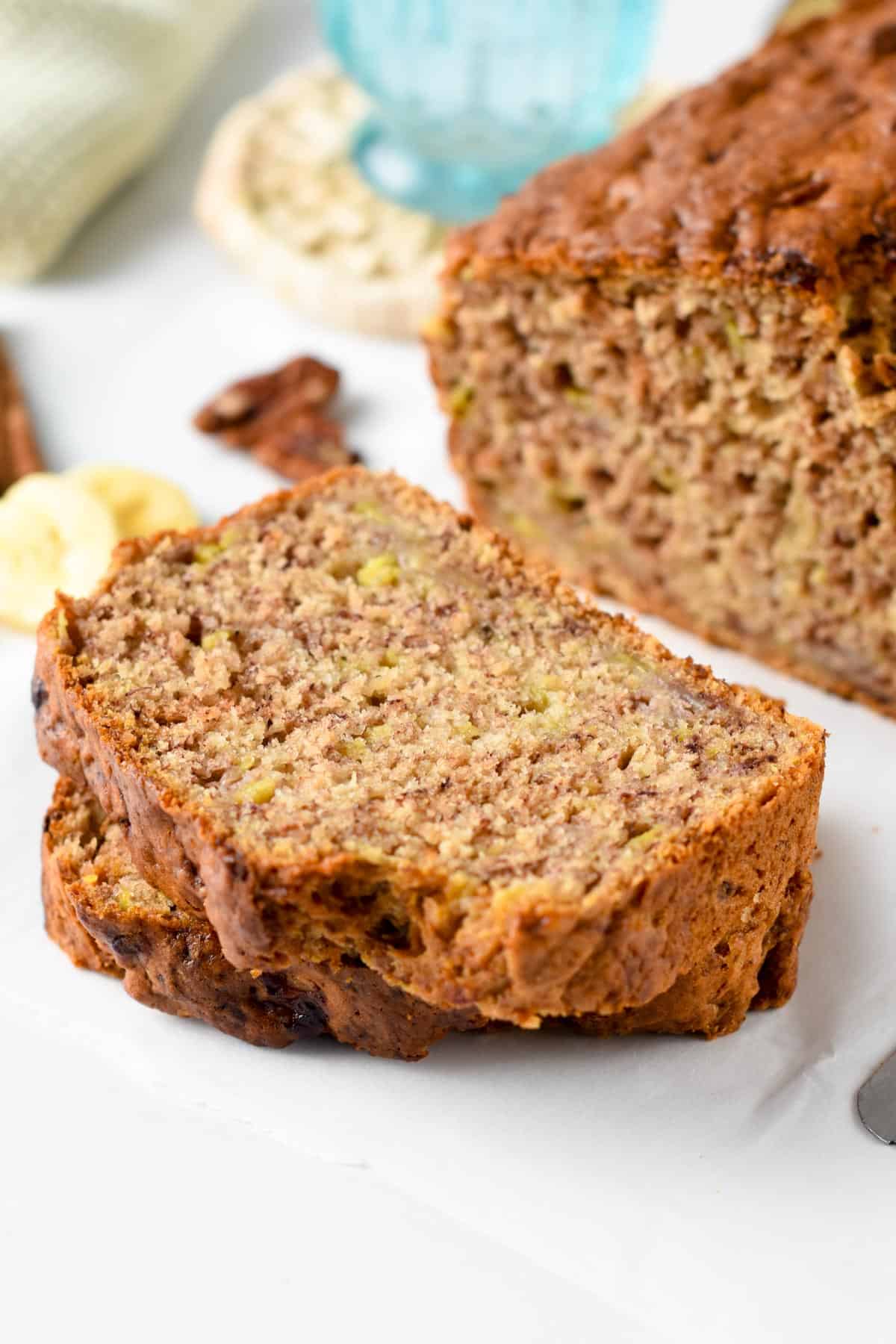 Two slices of Eggless Banana Bread flat on a plate in front of the whole loaf.