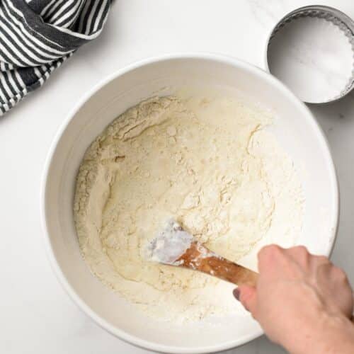 a mixing bowl filled with flour and liquids and a hand stirring the dough with a silicone spatula