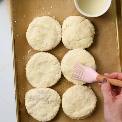a hand holding a pink silicone brush and brush the top of uncooked scones with coconut cream