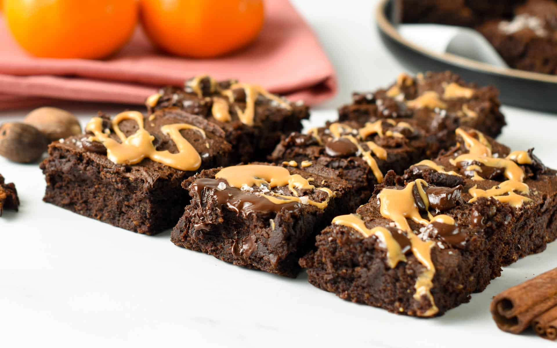 Pumpkin Brownies squares decorated with peanut butter, melted chocolate, and sea salt flakes in front of two oranges.