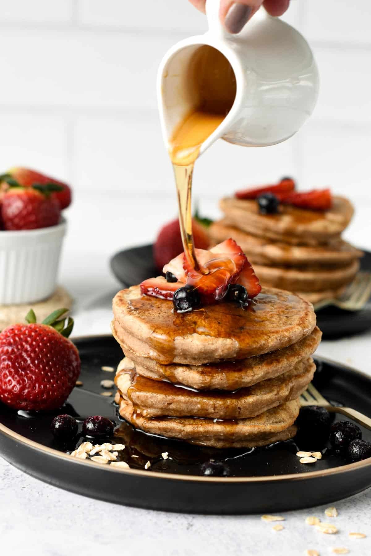 Pouring maple syrup on a stack of Vegan Oatmeal Pancakes on a black plate, decorated with sliced strawberries and blueberries.