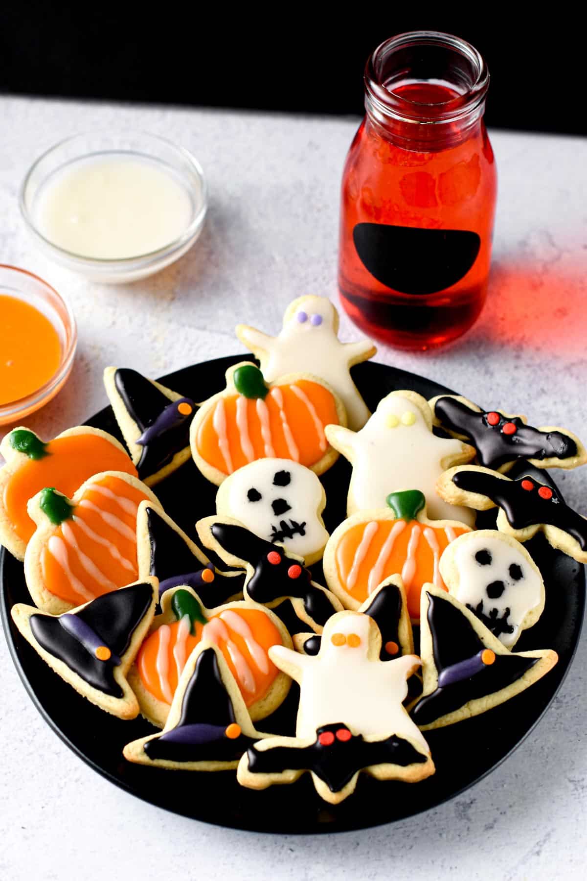Vegan Halloween Sugar Cookies in ghost, skull, pumpkin, witch hat, and bat shapes on a dark plate with a red drink in the background.
