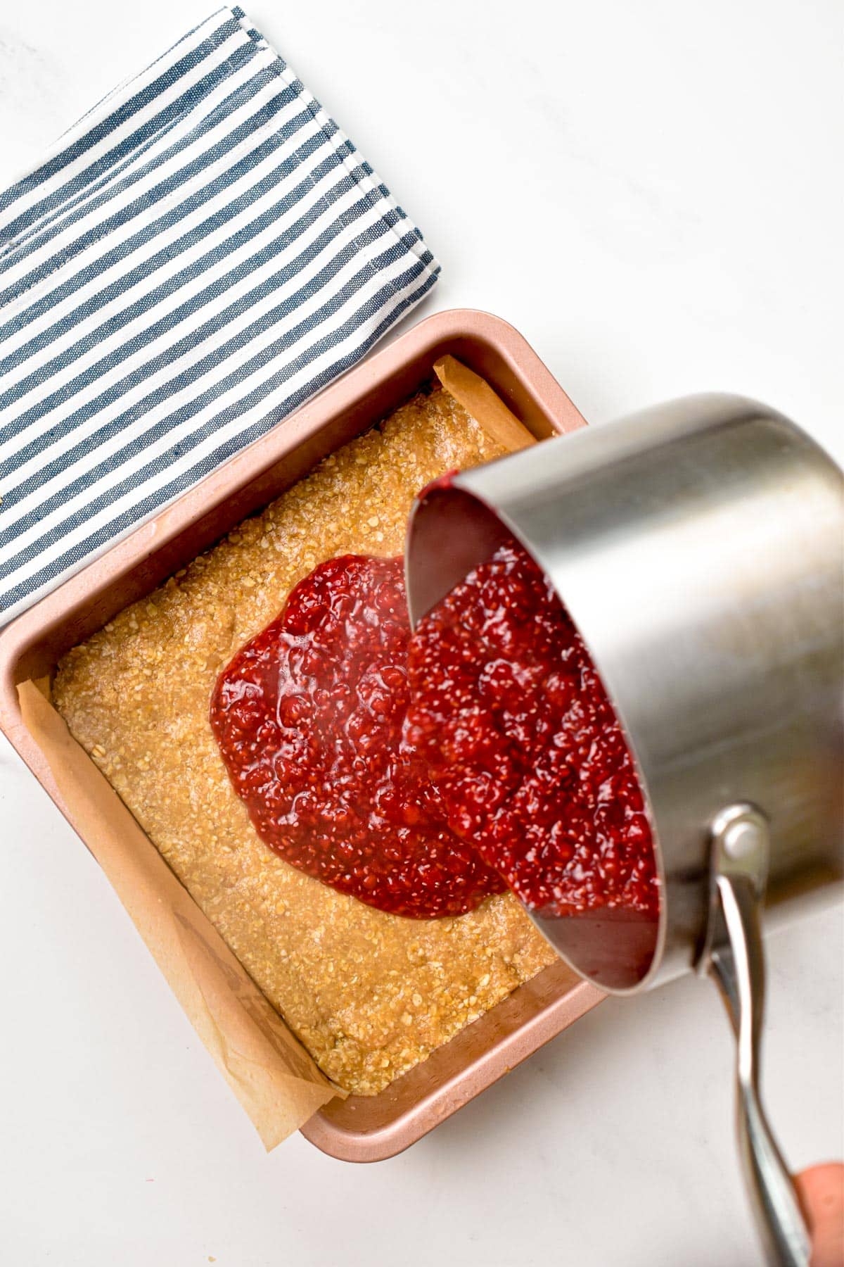 A saucepan pouring hot raspberry jam on top of the oatmeal bar mixture (unbaked).