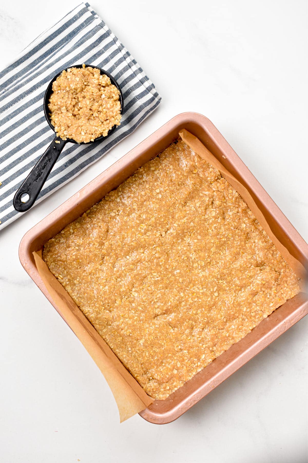 A square pan filled with a layer of oatmeal mixture and a 1/2 cup measuring cup filled with remaining batter.