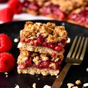 a stack of two Raspberry Oatmeal Bars filled with raspberry jam, on a black plate and with two fresh raspberries on the left side