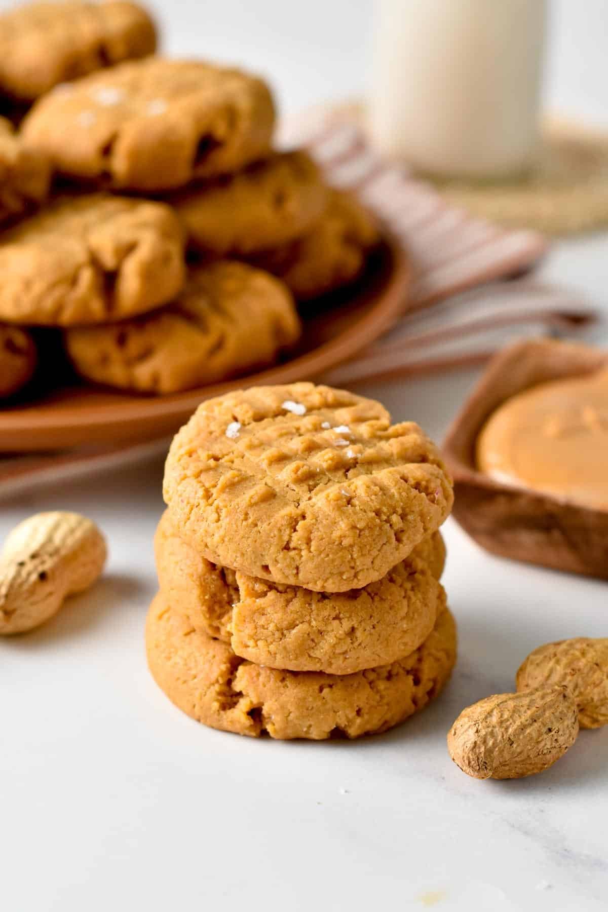 Vegan Peanut Butter Cookies stacked on a table in front of a bunch of them on a table.