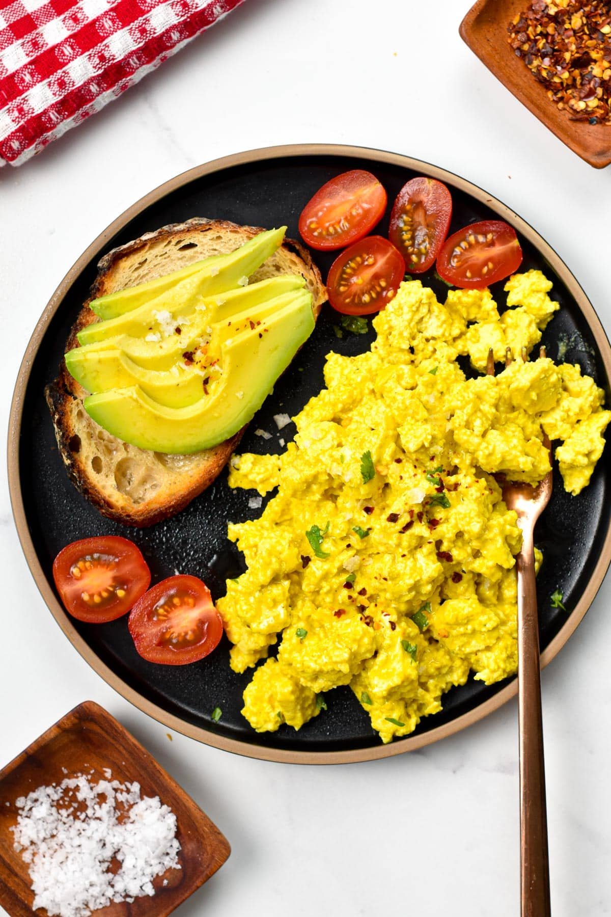 Tofu scramble on a black plate with an avocado on toast and sliced cherry tomatoes.