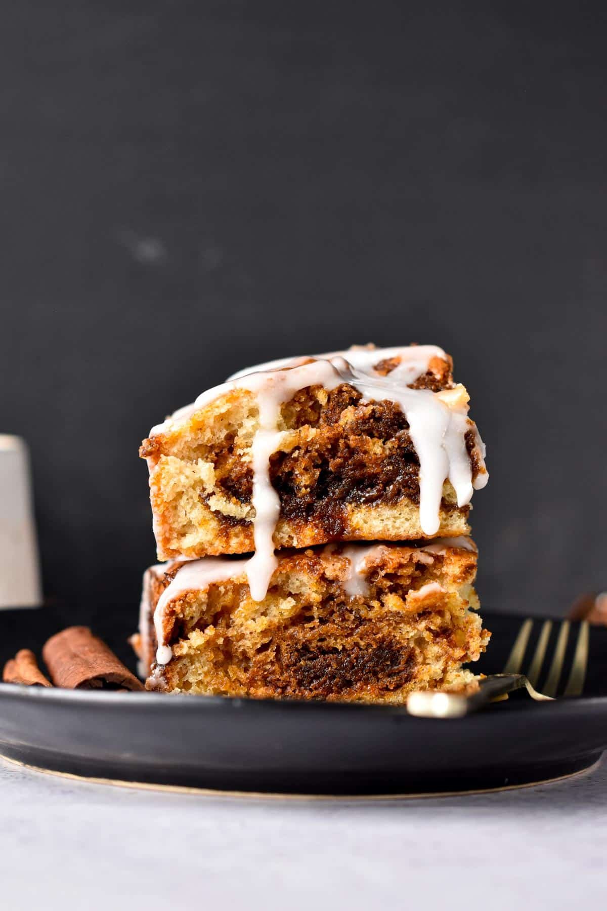 Two slices of Vegan Coffee Cake stacked on a black plate next to cinnamon sticks and a golden spoon.