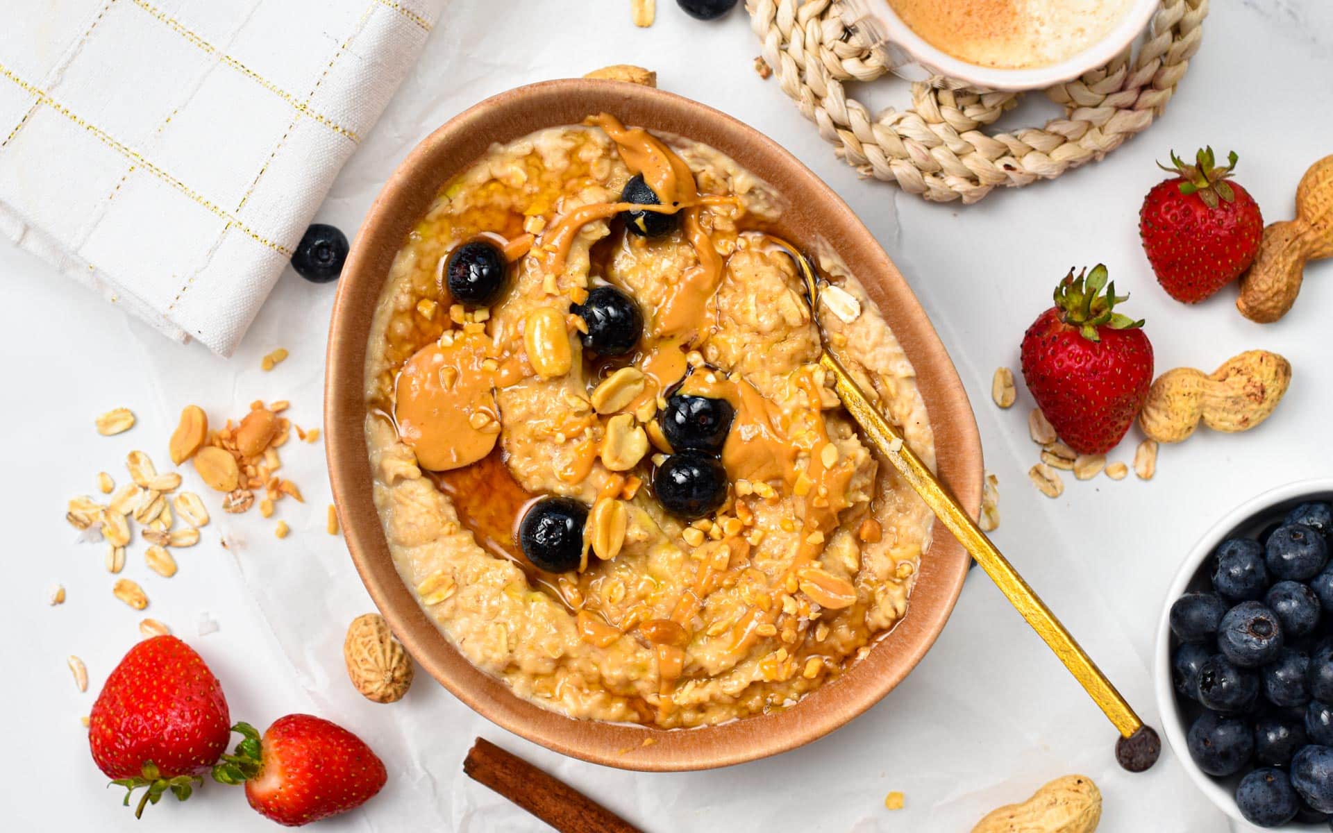 Peanut Butter Oatmeal bowl decorated with blueberries, with a golden spoon, next to fresh strawberries, blueberries, and peanuts.