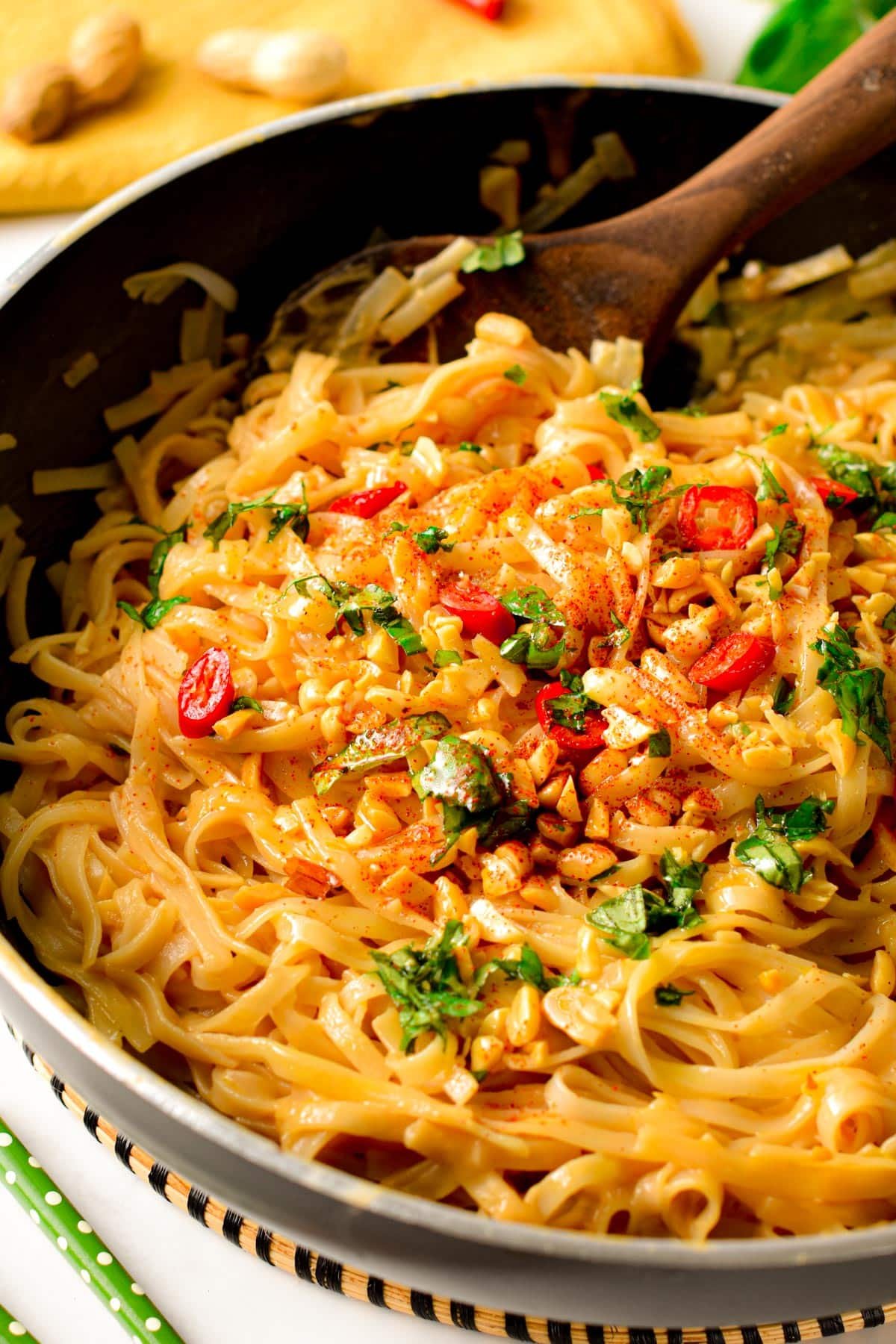 Peanut noodles in a large black bowl with a wooden spoon, decorated with Thai basil and chili slices.