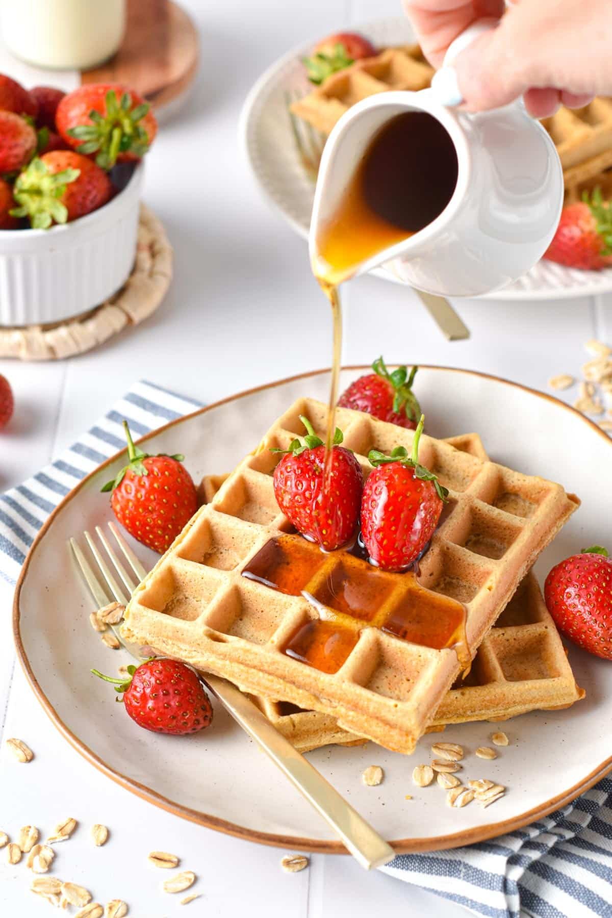 Pouring maple syrup on two waffles stacked on a plate full of strawberries.