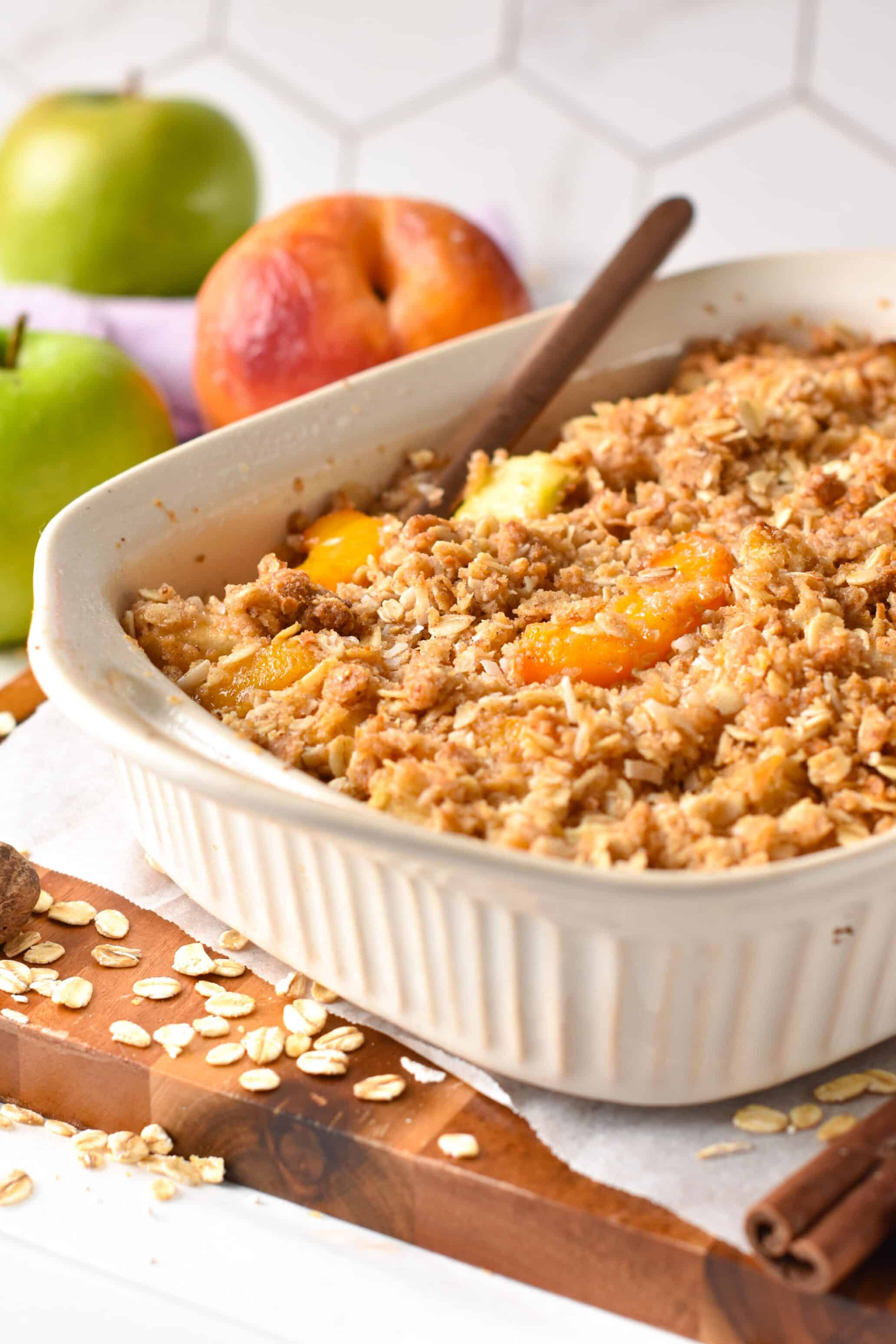 Apple and Peach Crumble in a white ceramic pan with a wooden spoon in it and apples next to it.
