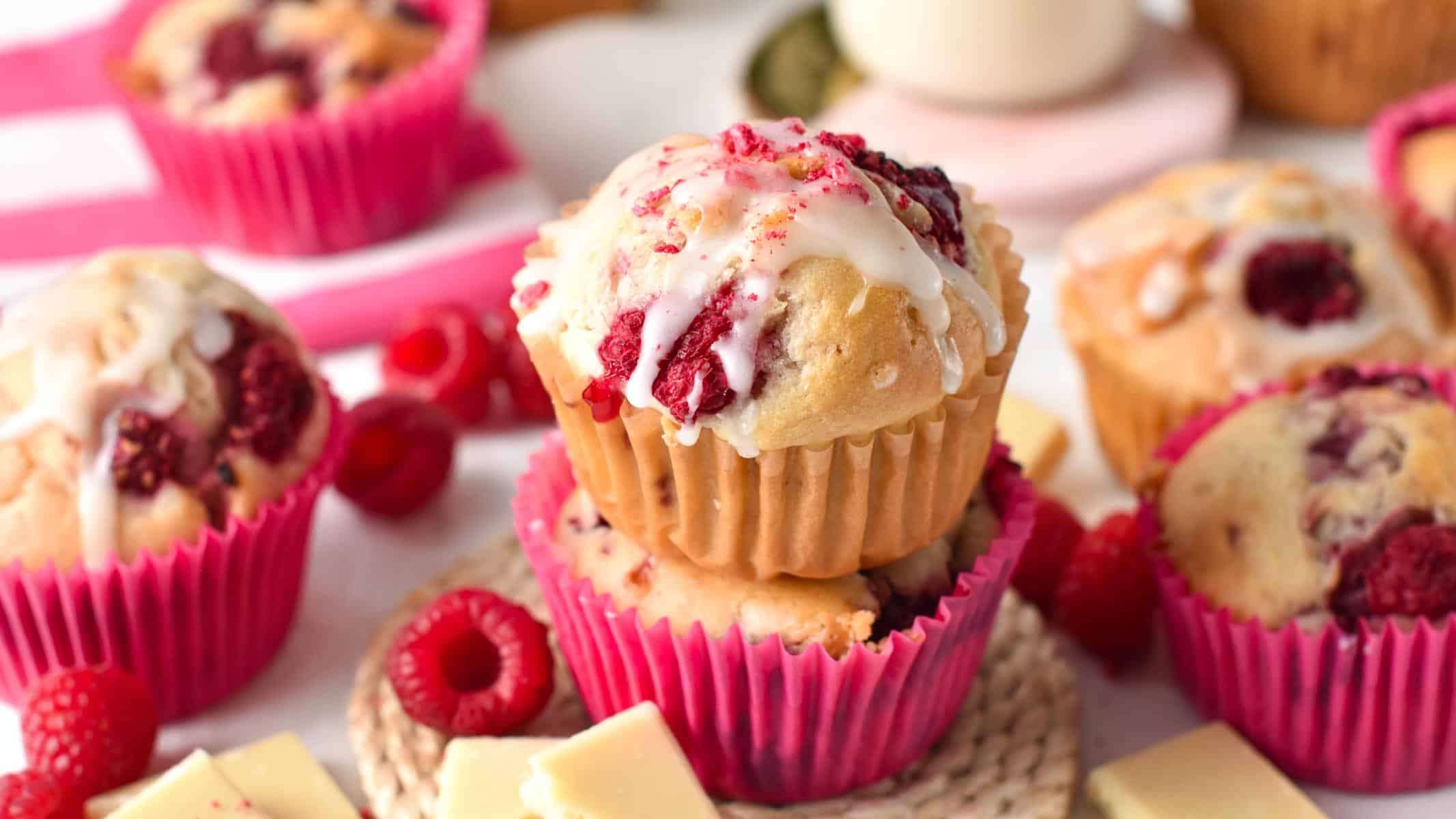 Vegan Raspberry Muffins stacked on a tabletop with their pink muffin case, decorated with fresh raspberries and melted vegan chocolate.